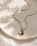 A gold snake chain necklace with a cherry pendant, resting on a light beige ceramic plate with soft leaf shadows. The pendant features a gold-toned metal setting with a clear pavé stone, a deep red gemstone, and a small green stone.