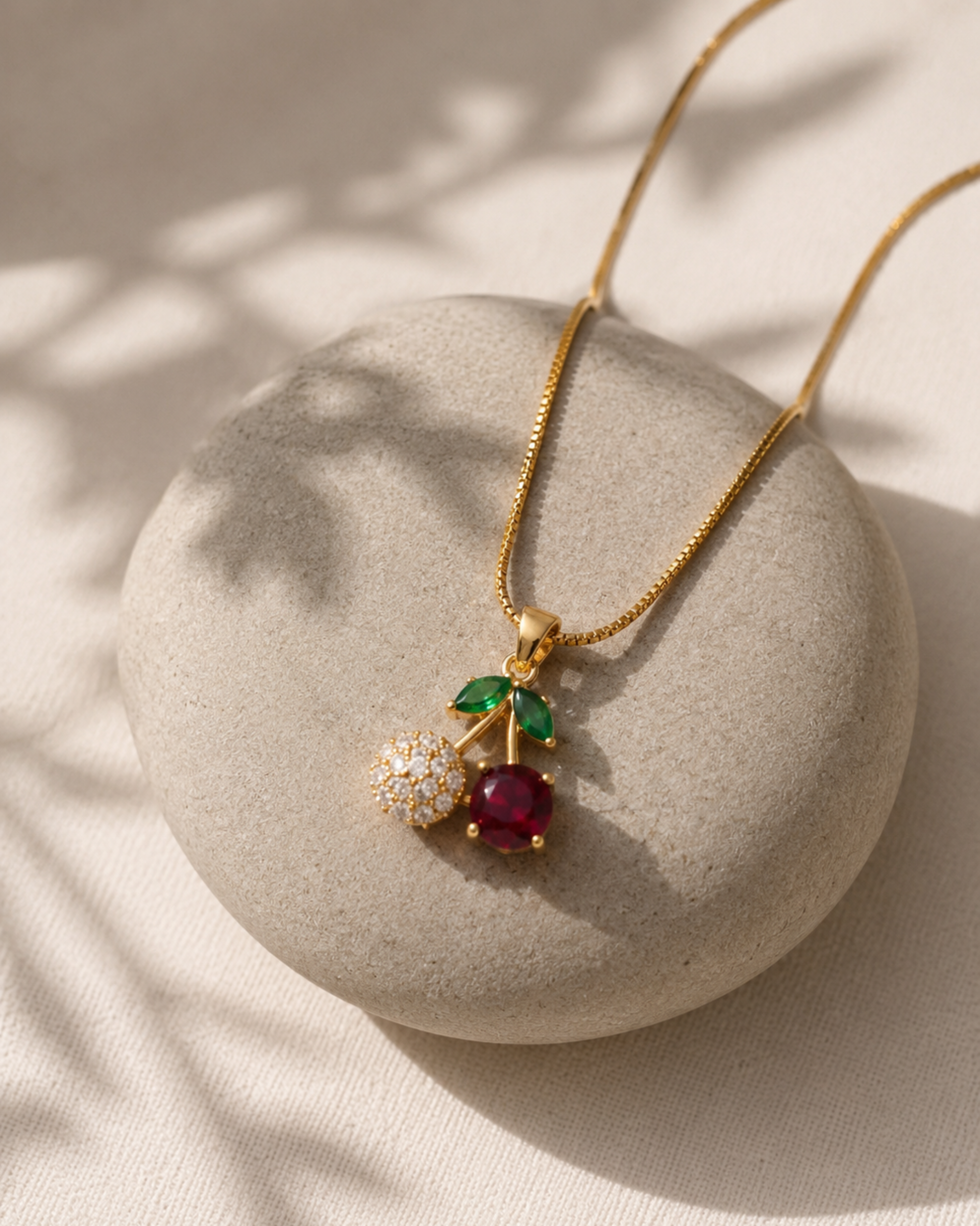 A gold chain necklace with a cherry pendant resting on a round beige stone. The pendant features a red gemstone cherry, a green leaf, and a white gemstone-covered bud, set against a softly lit beige background with leaf shadows.
