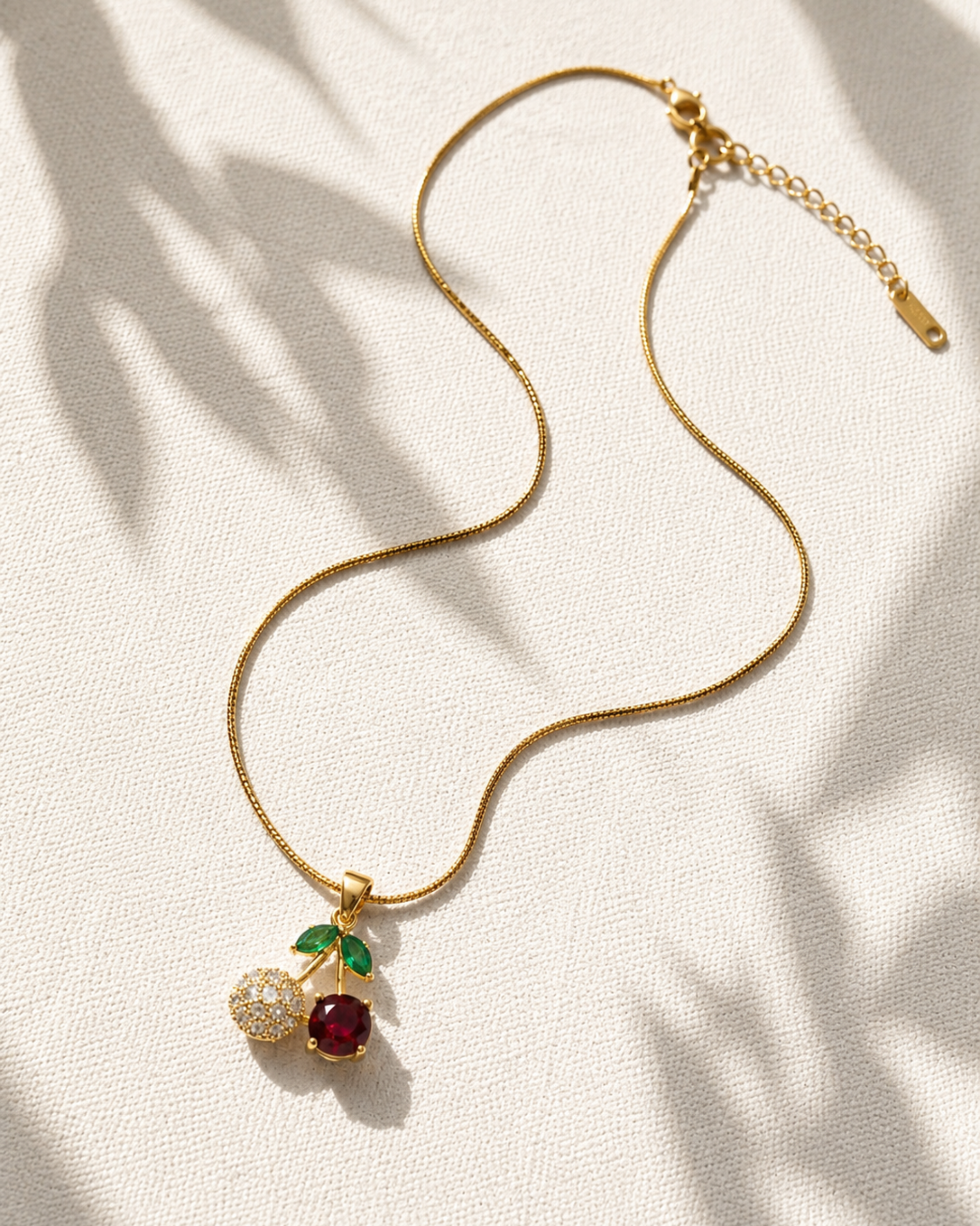 A gold chain necklace with a cherry pendant, featuring a red gemstone, a clear crystal-studded half cherry, and green leaf accents, displayed on a textured cream fabric with soft palm leaf shadows.