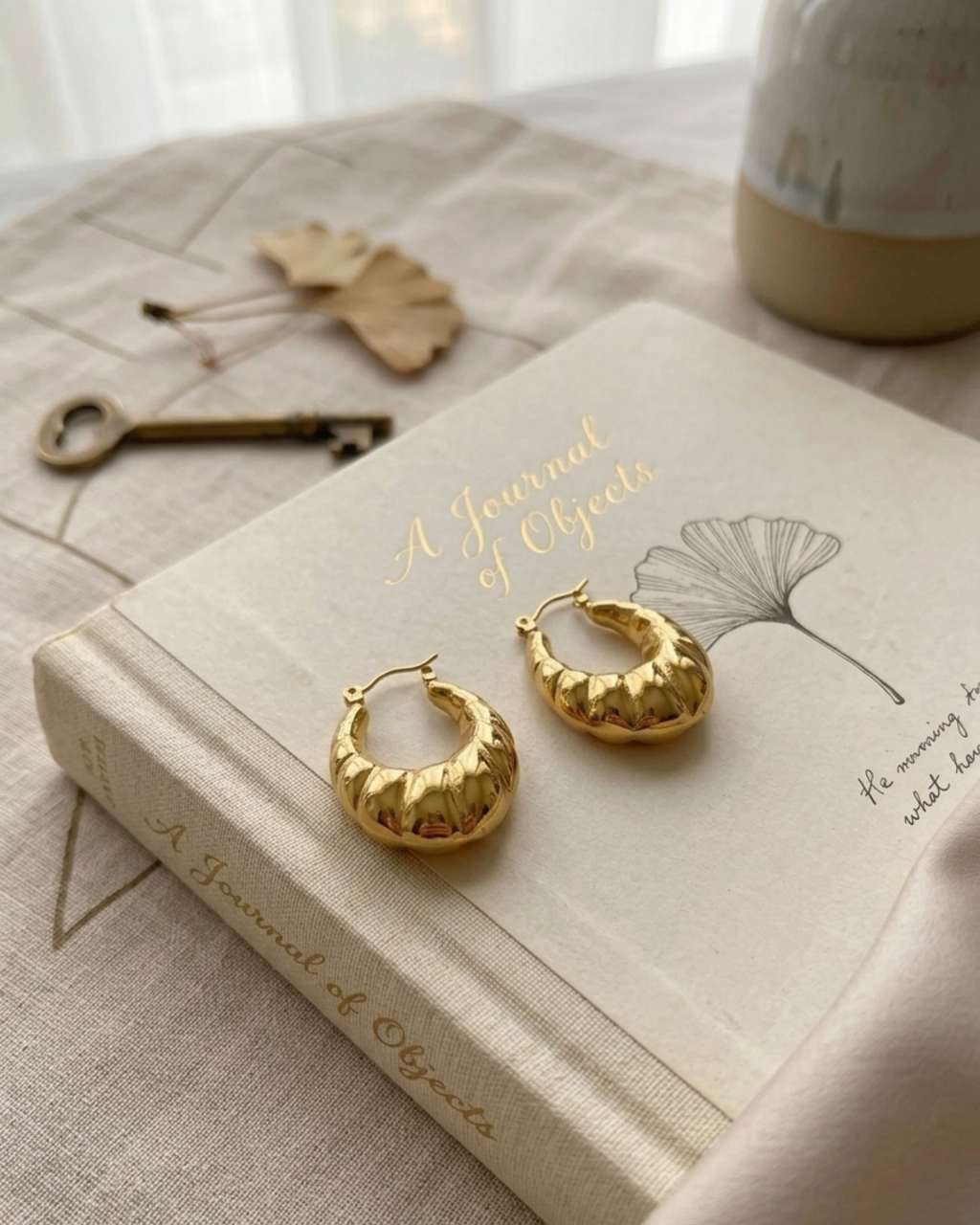 A pair of textured gold hoop earrings displayed on a book titled 'A Journal of Objects' on a beige fabric surface with a vintage key and dried leaves nearby.