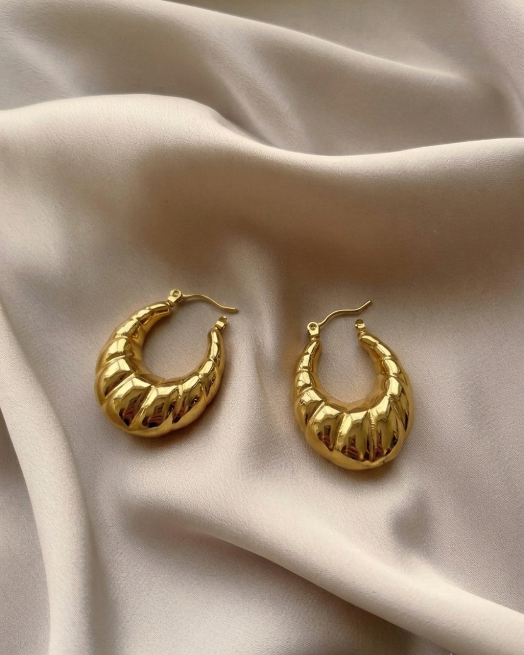 A pair of textured gold crescent hoop earrings resting on draped cream-colored satin fabric.