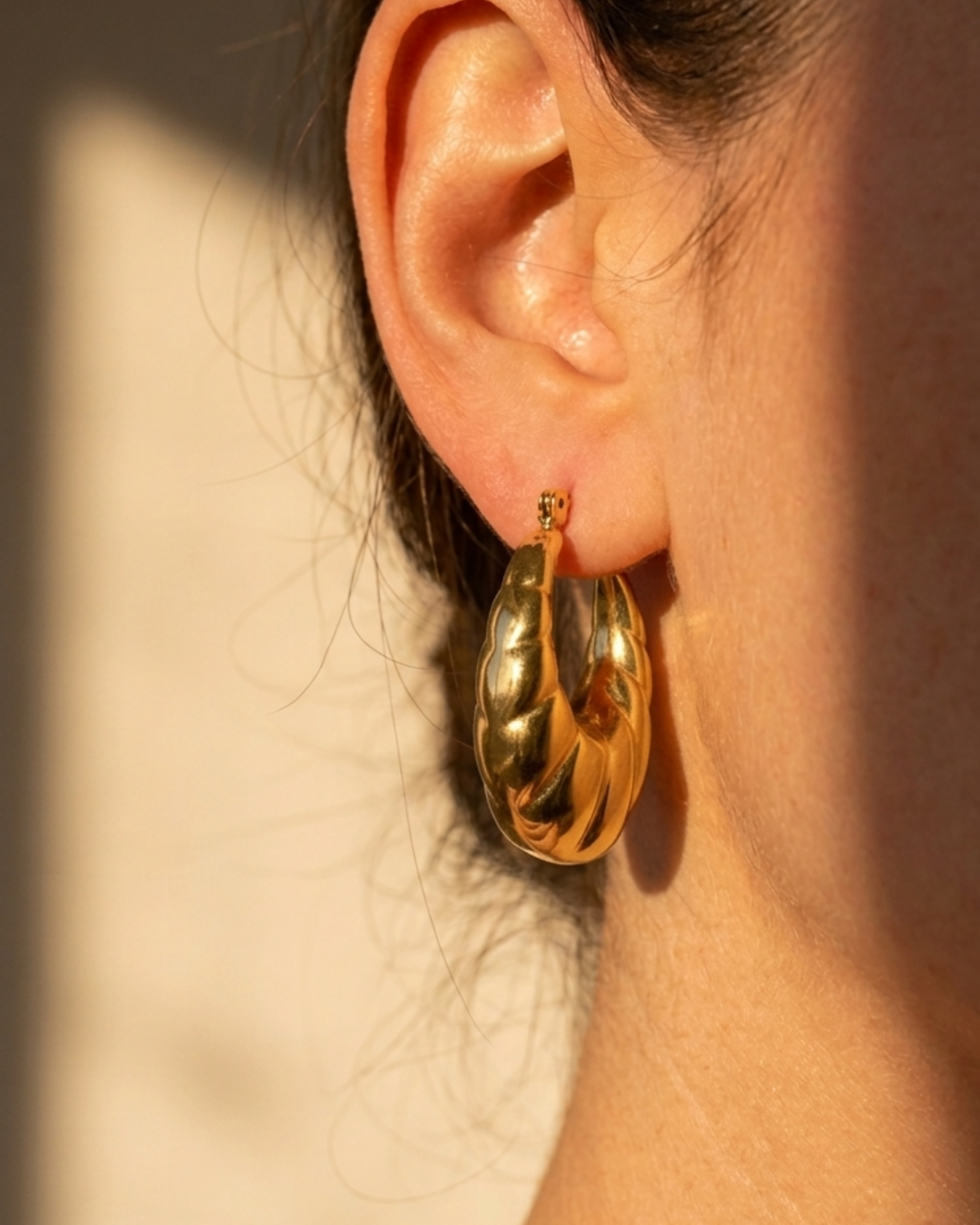 A close-up of a person's ear wearing a chunky, polished gold hoop earring, with warm sunlight casting shadows on the skin and background.