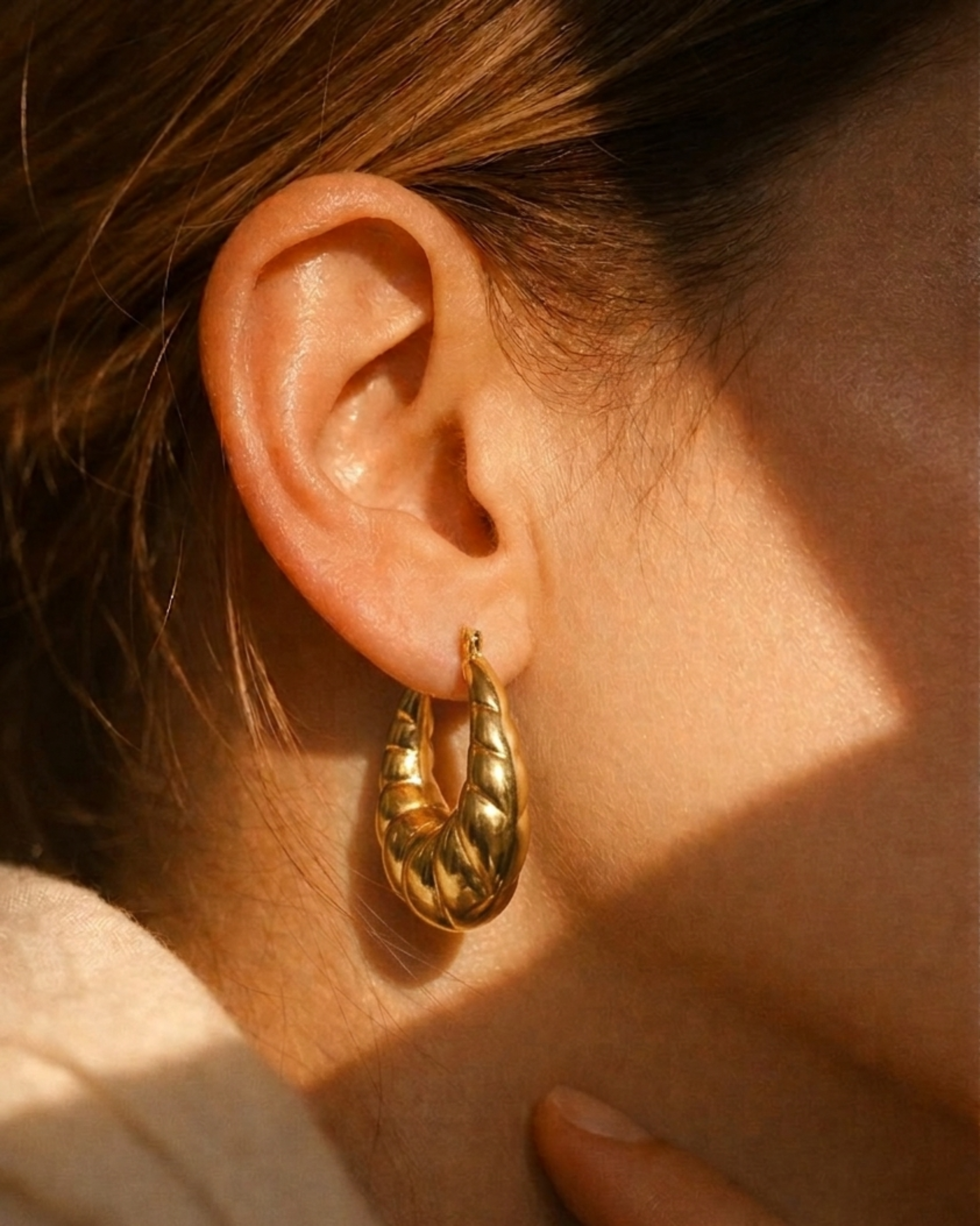A close-up of a person's ear wearing a textured gold hoop earring, with warm sunlight casting shadows on the skin and hair.