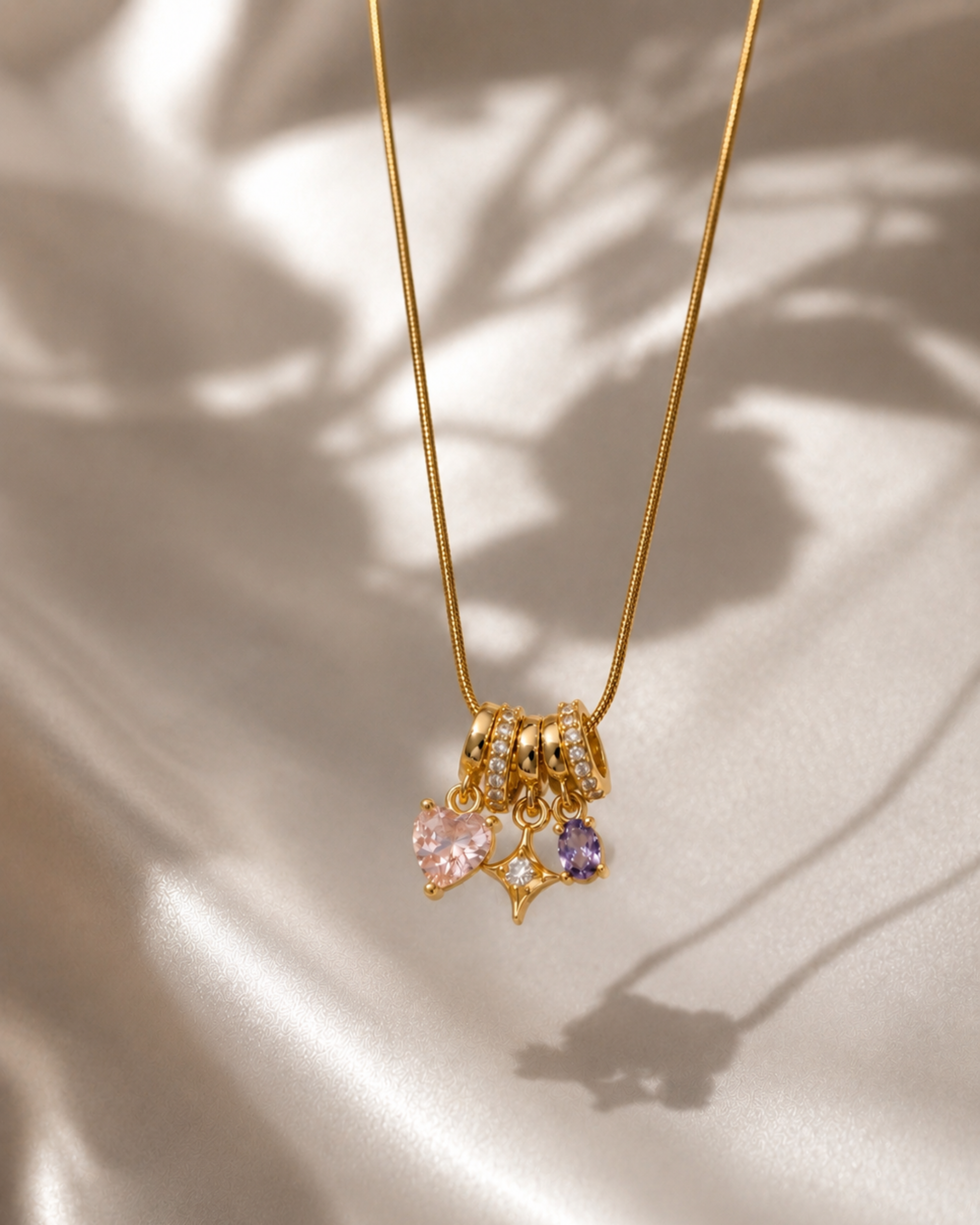 A gold chain necklace with a cluster of charms, including a pink heart-shaped crystal, a purple star-shaped crystal, and textured gold rings, resting on a draped champagne satin fabric with soft leaf shadows.