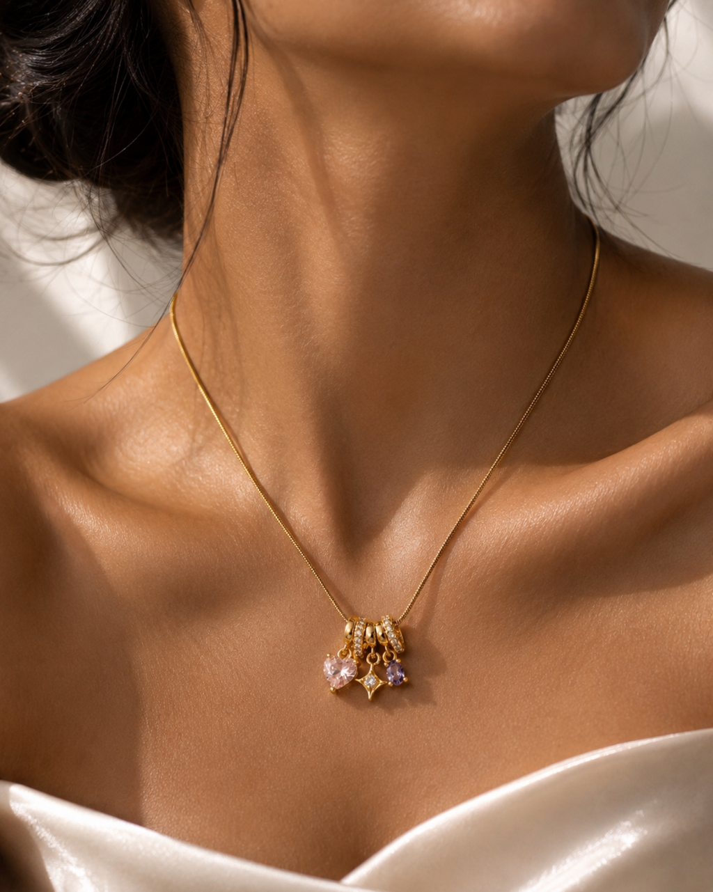 A close-up of a model's neck and chest wearing a delicate gold chain necklace with a small cluster pendant featuring light pink and lavender gemstones.