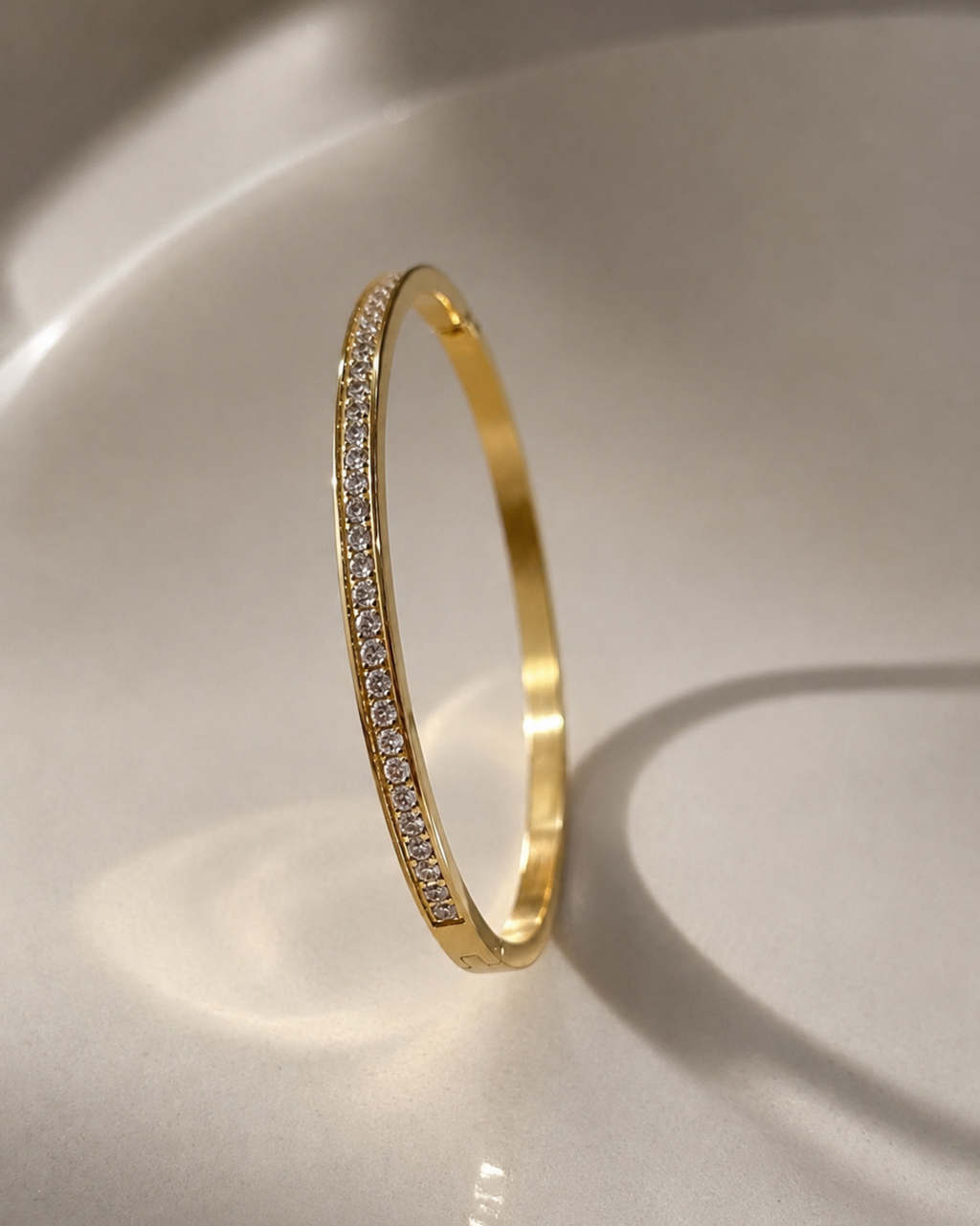 A slim gold bangle bracelet with a single row of small clear pavé stones along one side, standing upright on a glossy white surface.