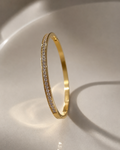 A slim gold bangle bracelet with a single row of small clear pavé stones along one side, standing upright on a glossy white surface.