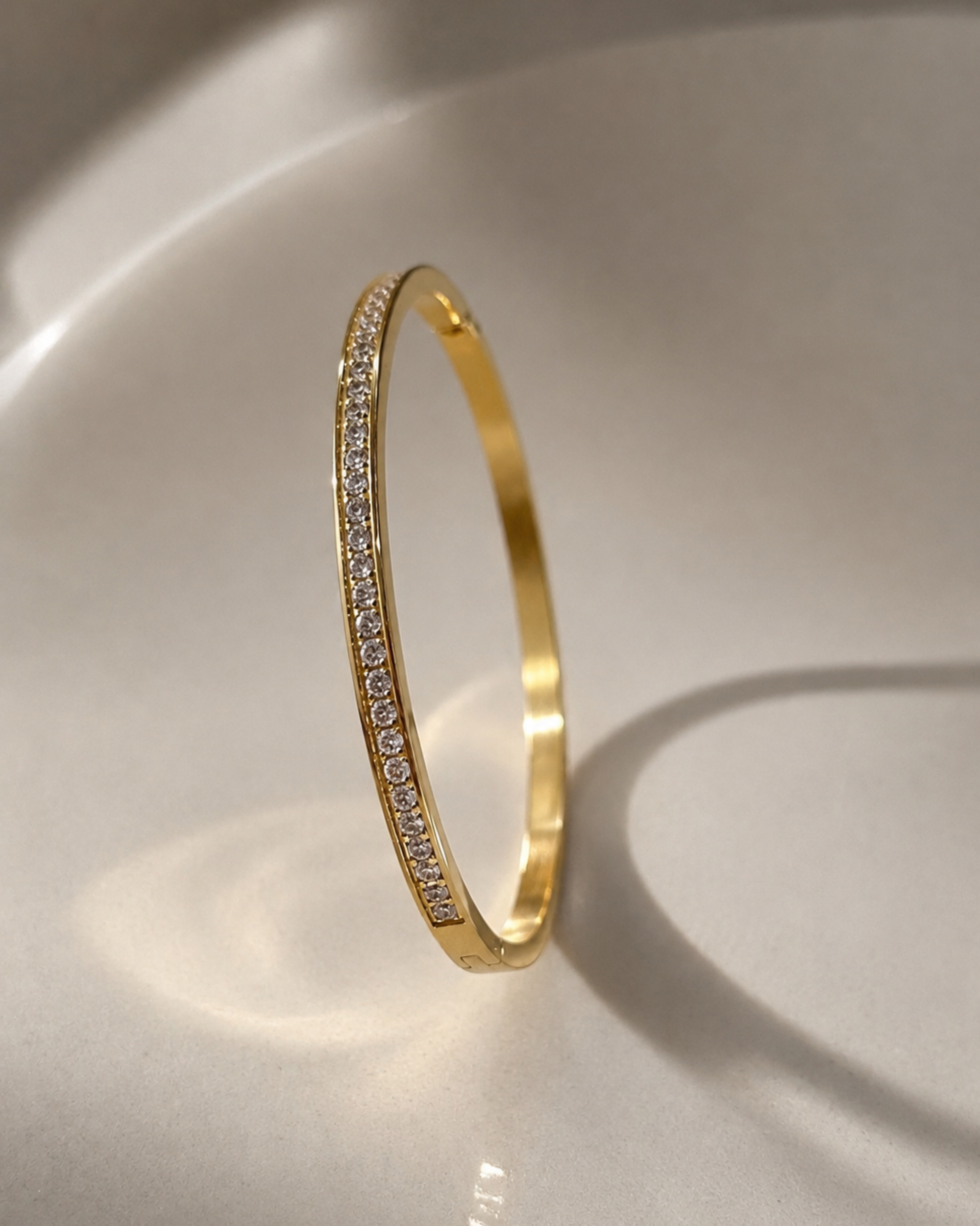 A slim gold bangle bracelet with a single row of small clear pavé stones along one side, standing upright on a glossy white surface.