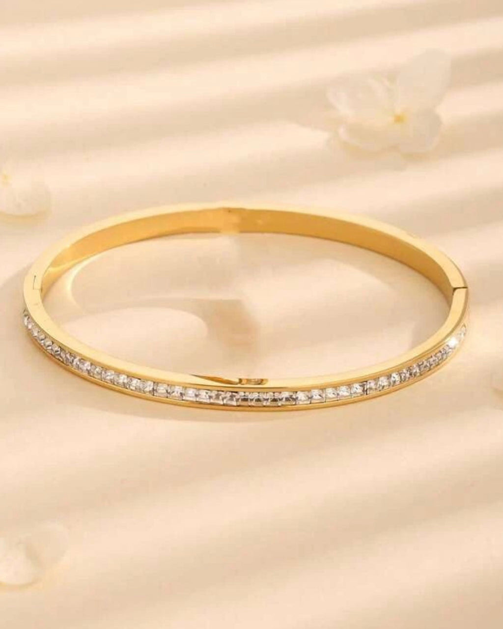 A gold hinged bangle bracelet with a row of small clear stones along the front band, displayed on a soft beige textured surface with a blurred flower in the background.