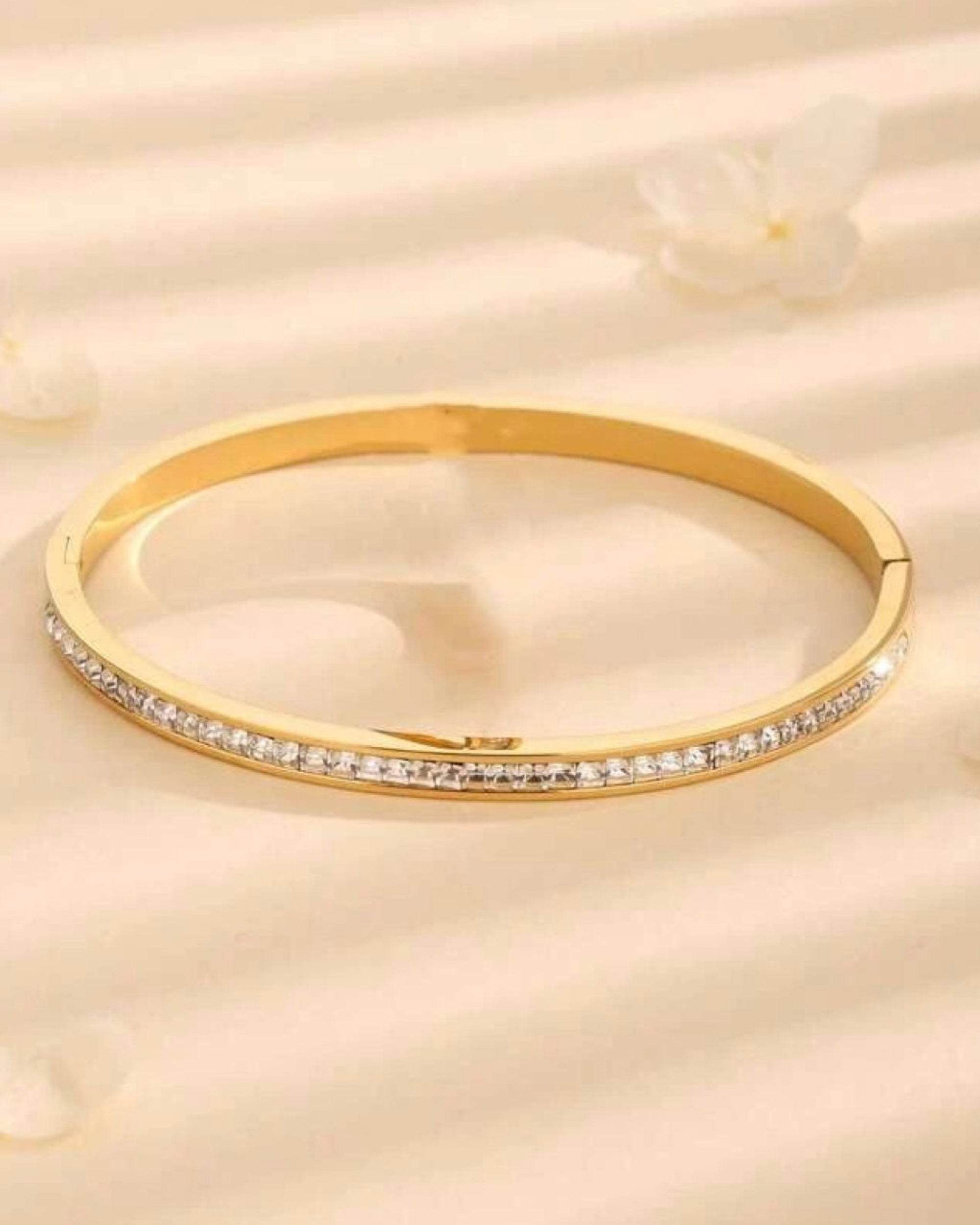 A gold hinged bangle bracelet with a row of small clear stones along the front band, displayed on a soft beige textured surface with a blurred flower in the background.