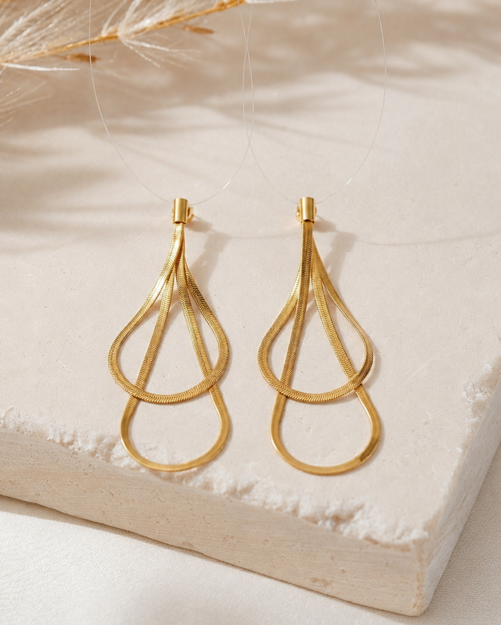 A pair of gold teardrop-shaped dangling earrings with textured inner loops and smooth outer loops, displayed on a light beige stone slab with soft dried grass shadows.