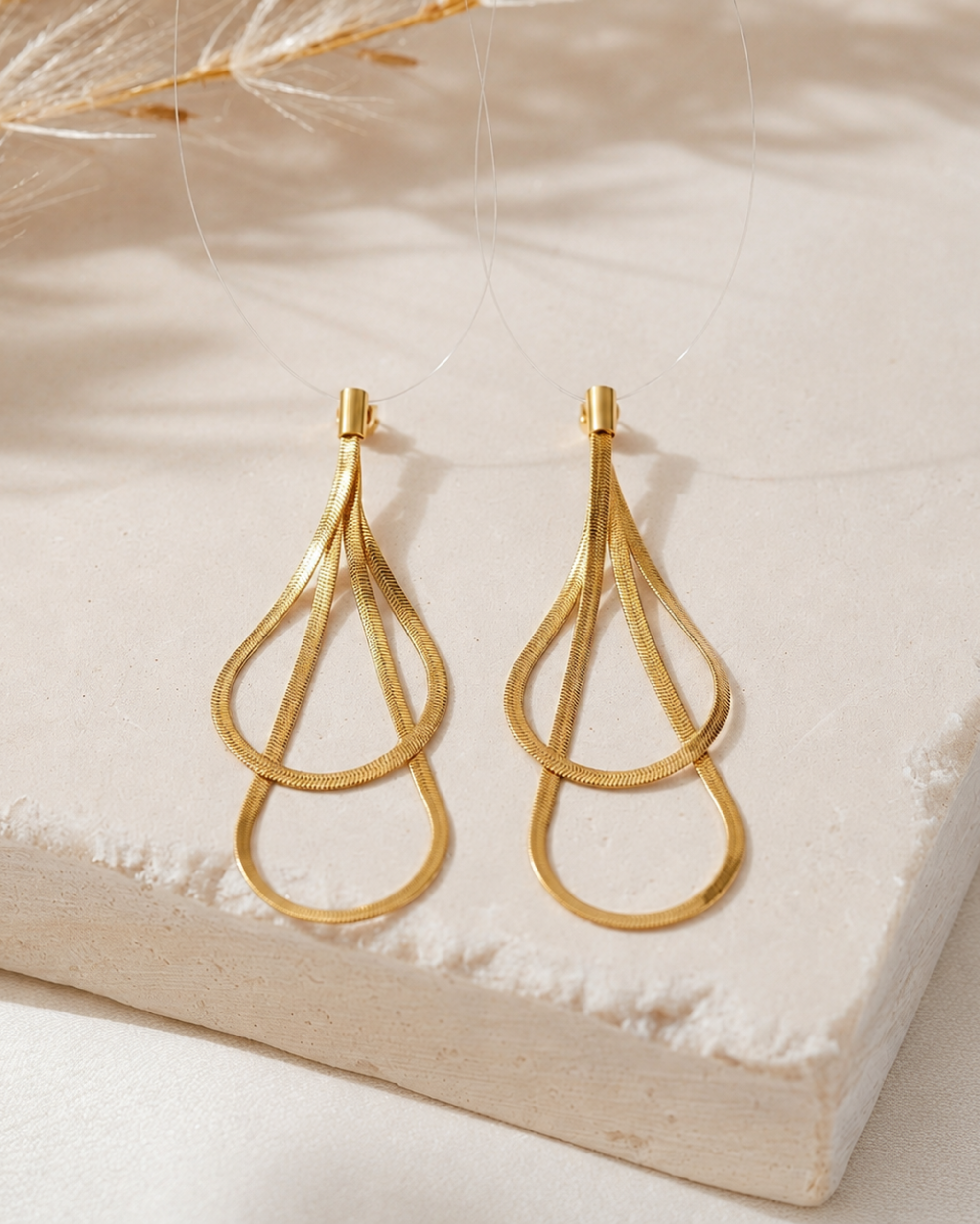 A pair of gold teardrop-shaped dangling earrings with textured inner loops and smooth outer loops, displayed on a light beige stone slab with soft dried grass shadows.