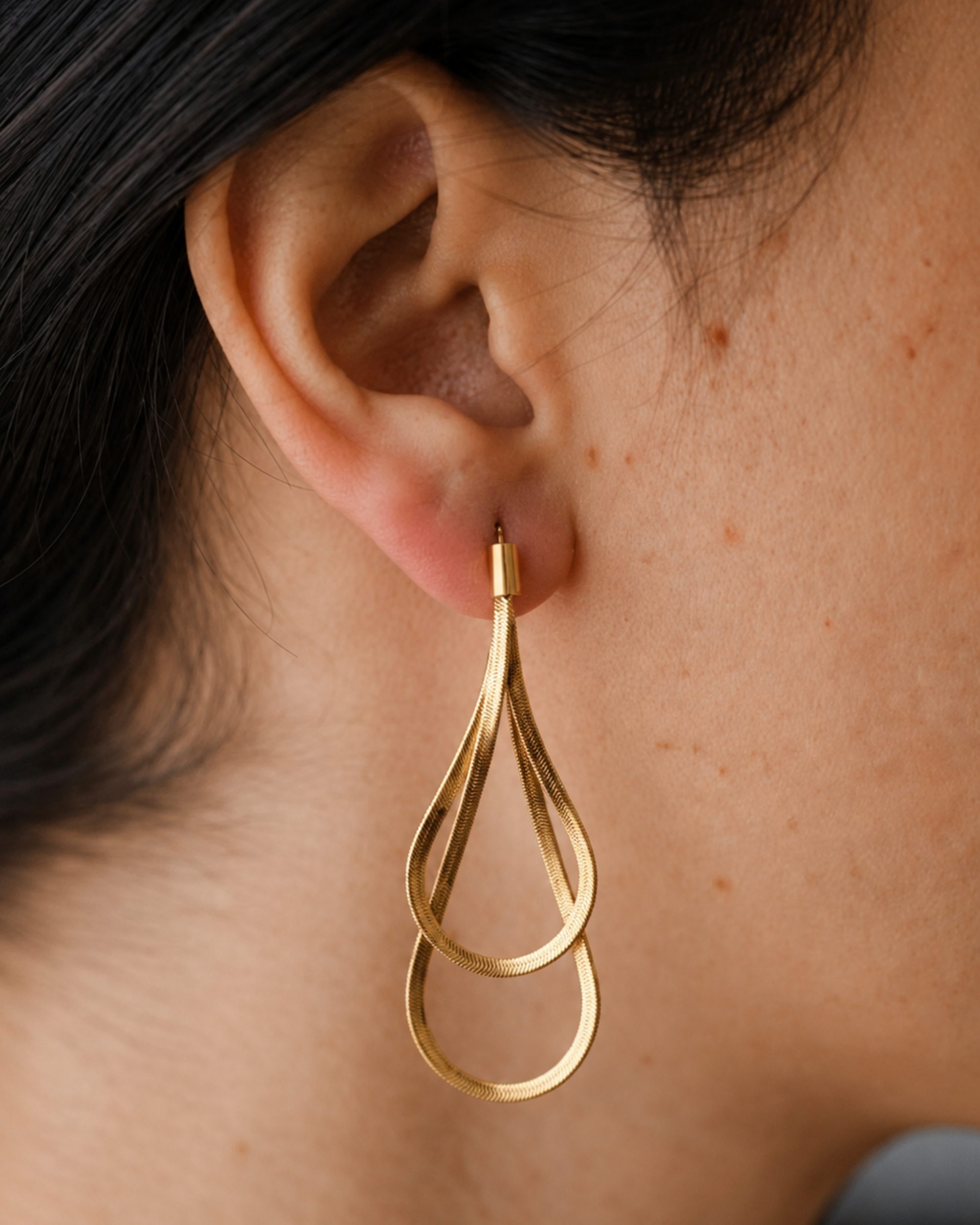 A close-up of a person's ear wearing a gold teardrop-shaped dangle earring with two interlocking loops.