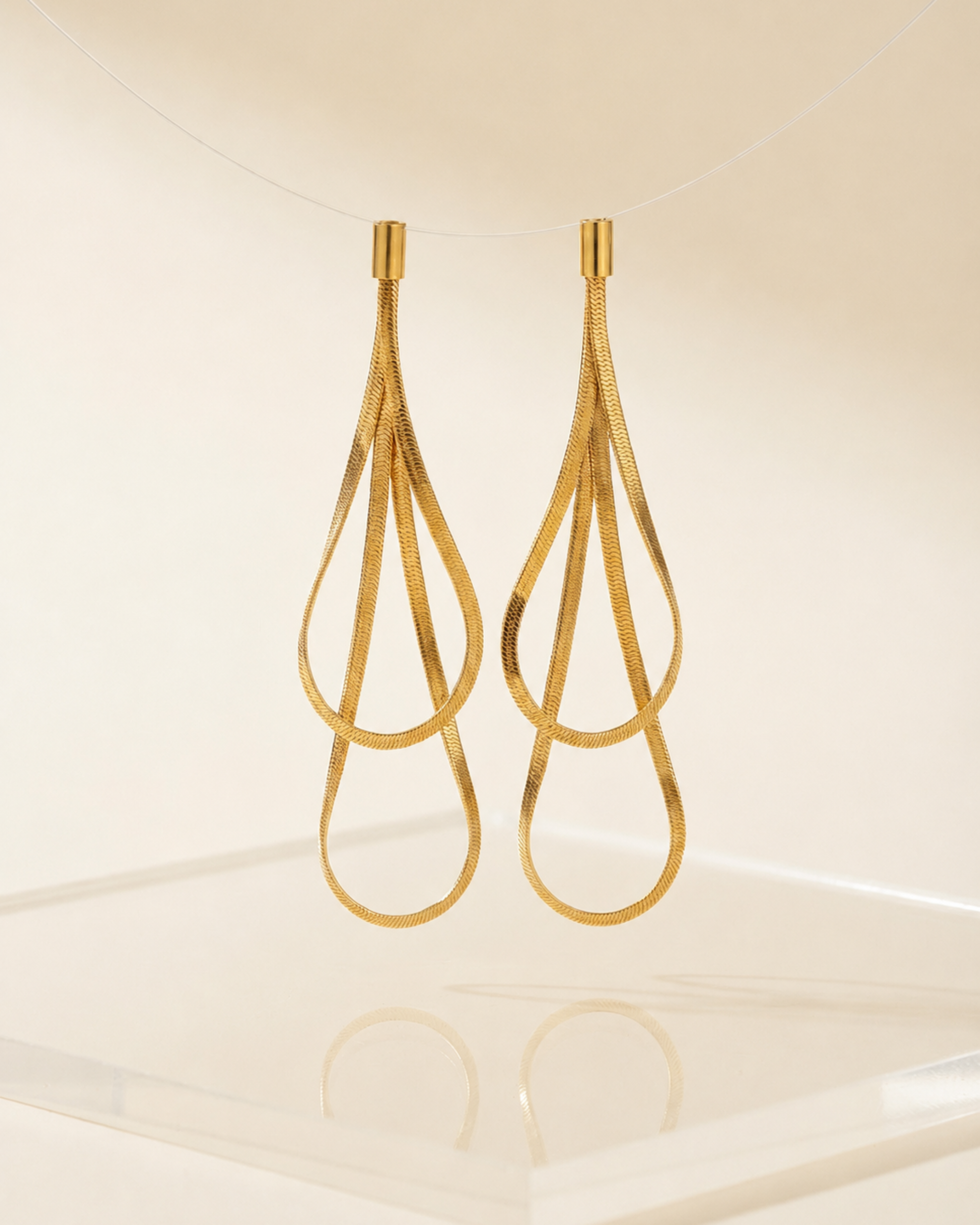 A pair of gold teardrop-shaped dangle earrings with a textured, snake-like woven design, hanging against a warm cream background.