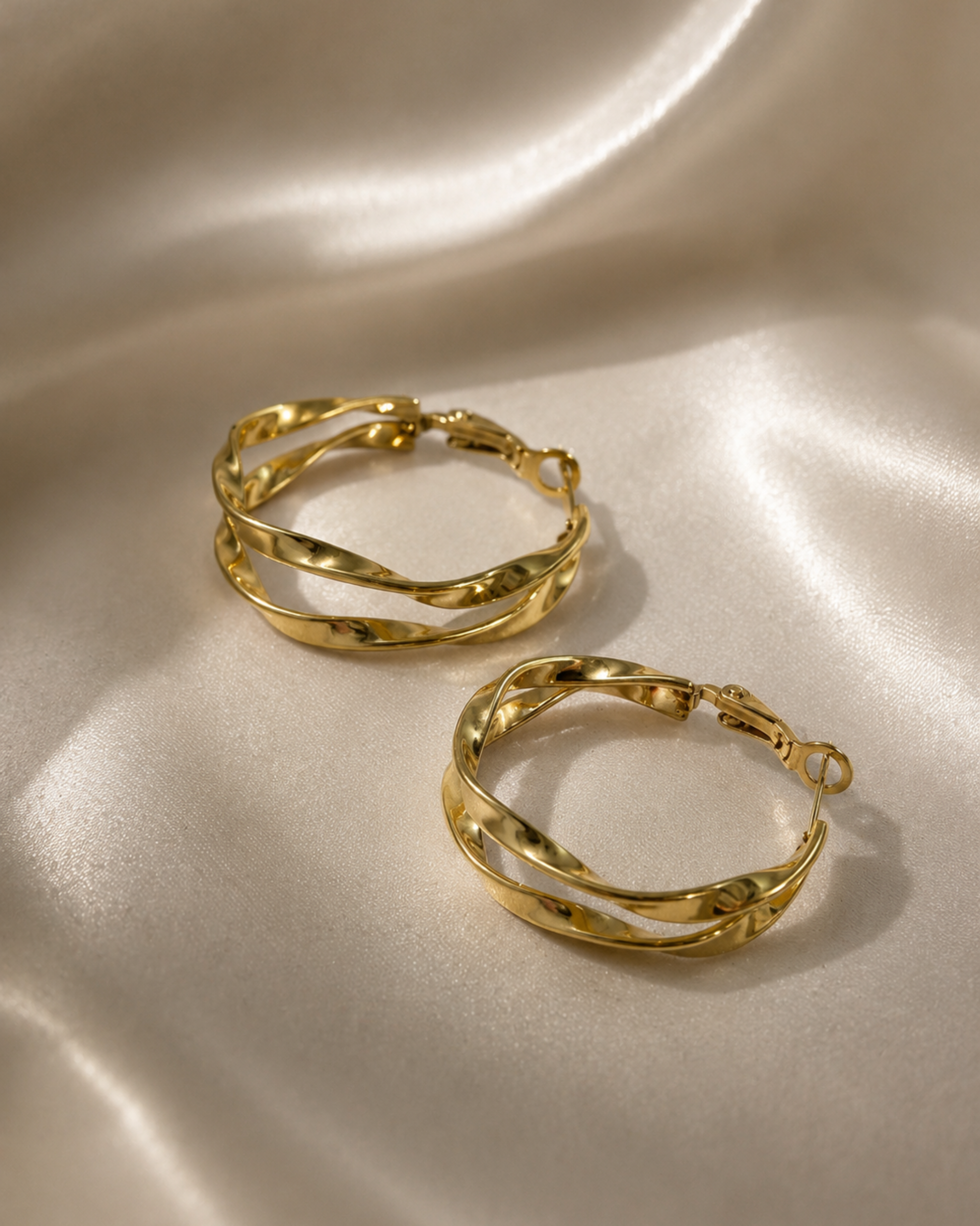 A pair of gold hoop earrings with a twisted, wavy design, displayed on a draped satin fabric background.