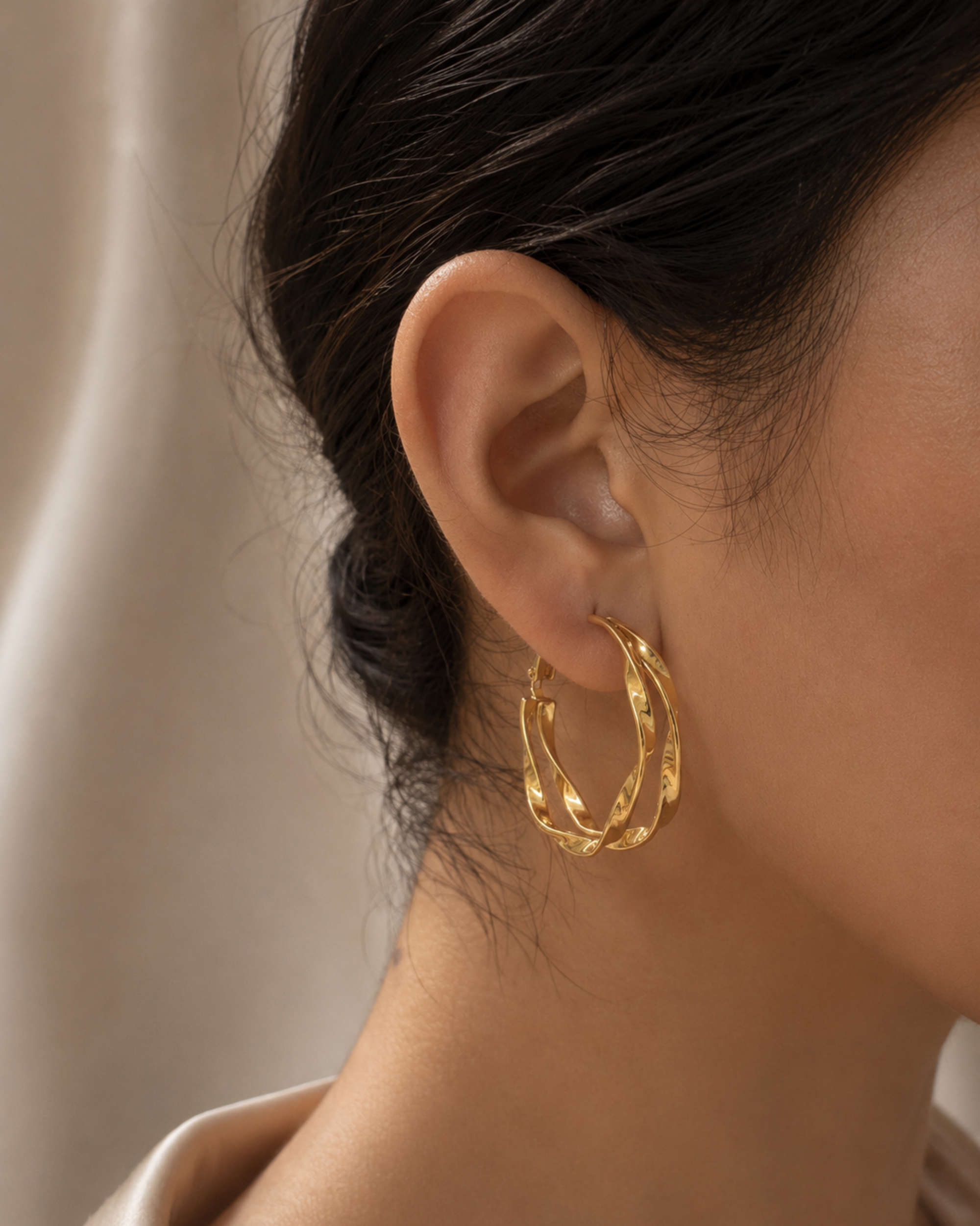 A close-up of a model's ear wearing a pair of textured gold hoop earrings against a soft beige background.