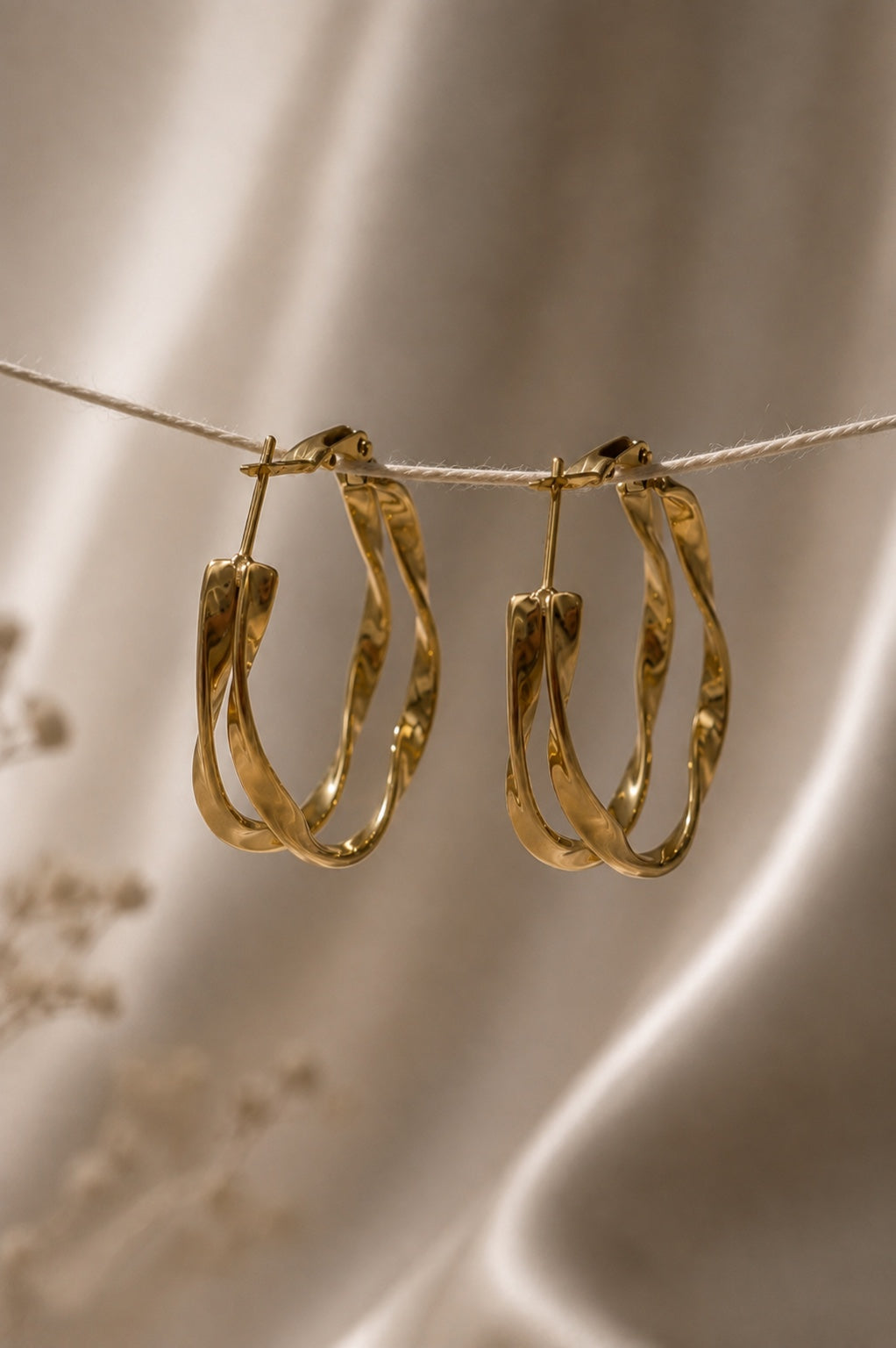 A pair of textured gold hoop earrings hanging from a thin string against a soft beige satin fabric background.