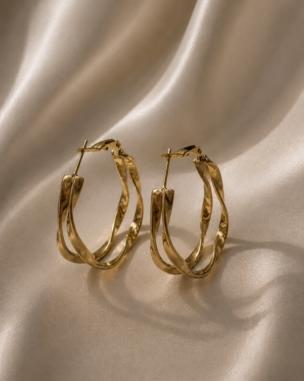 A pair of gold hoop earrings with a textured, twisted design, displayed on a draped beige satin fabric background.