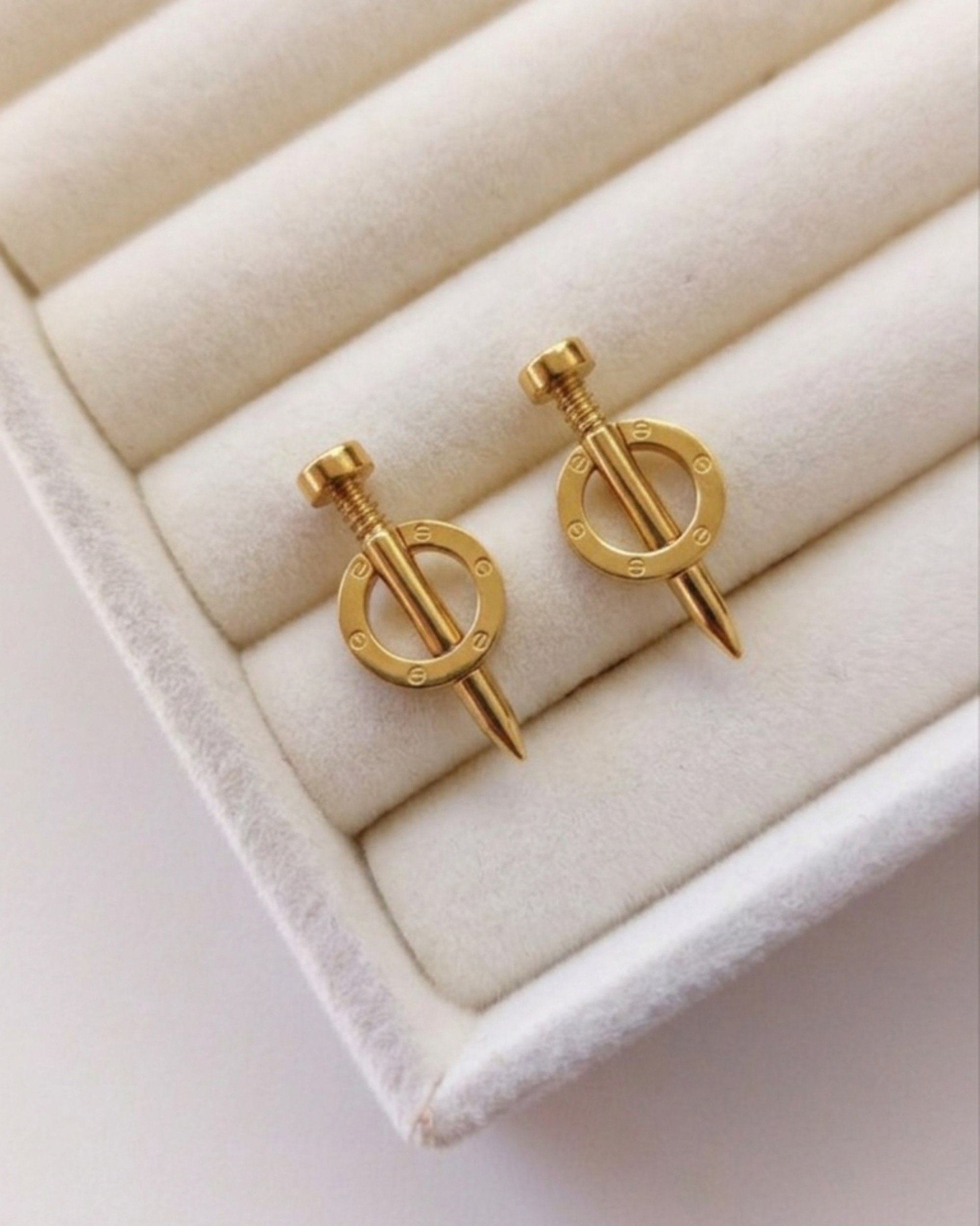 A pair of gold earrings with a circular pendant and a small pointed pin below, displayed on a cream-colored jewelry box cushion.