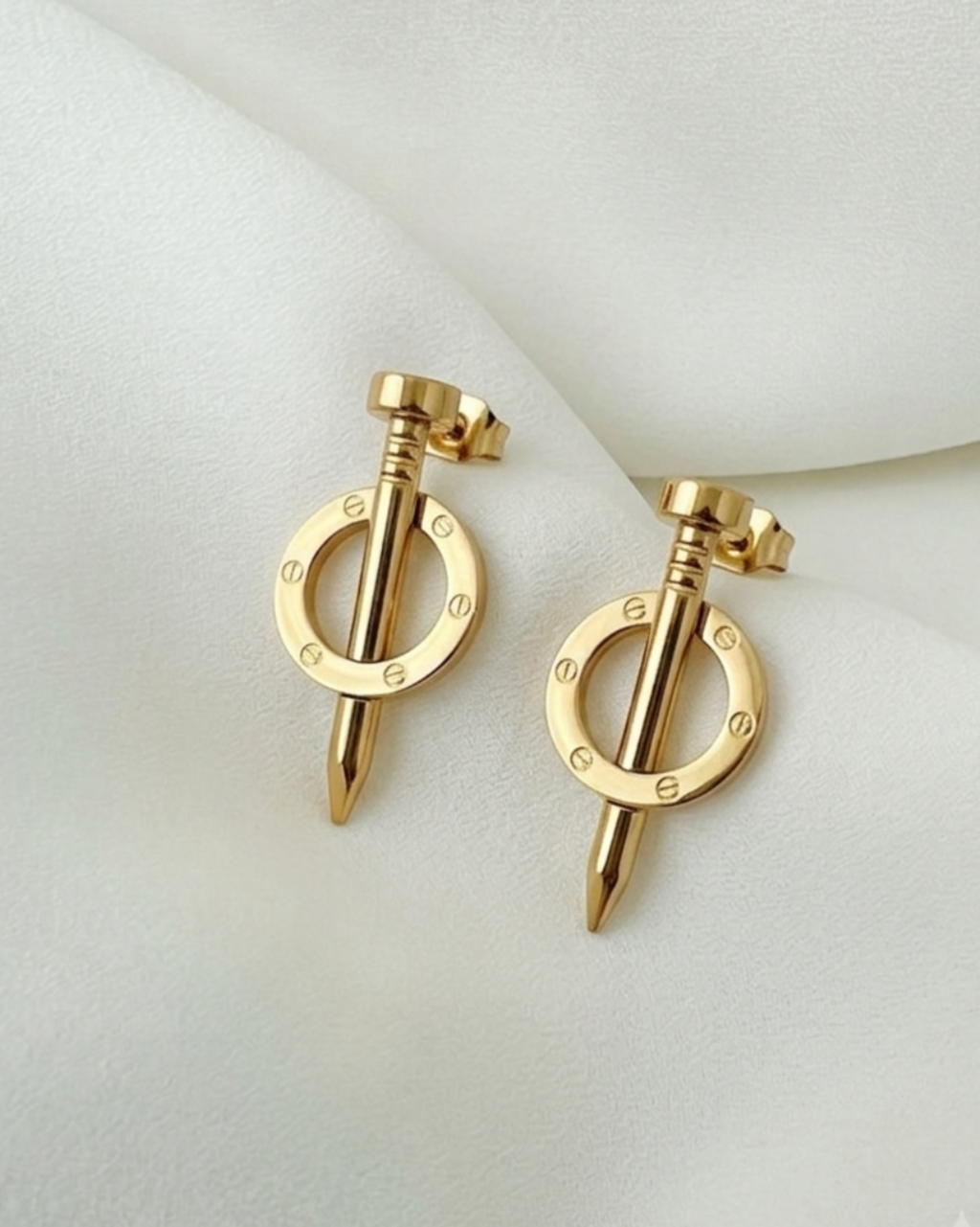A pair of gold-toned hoop earrings with a central spike and small engraved letters, displayed on a soft white fabric background.