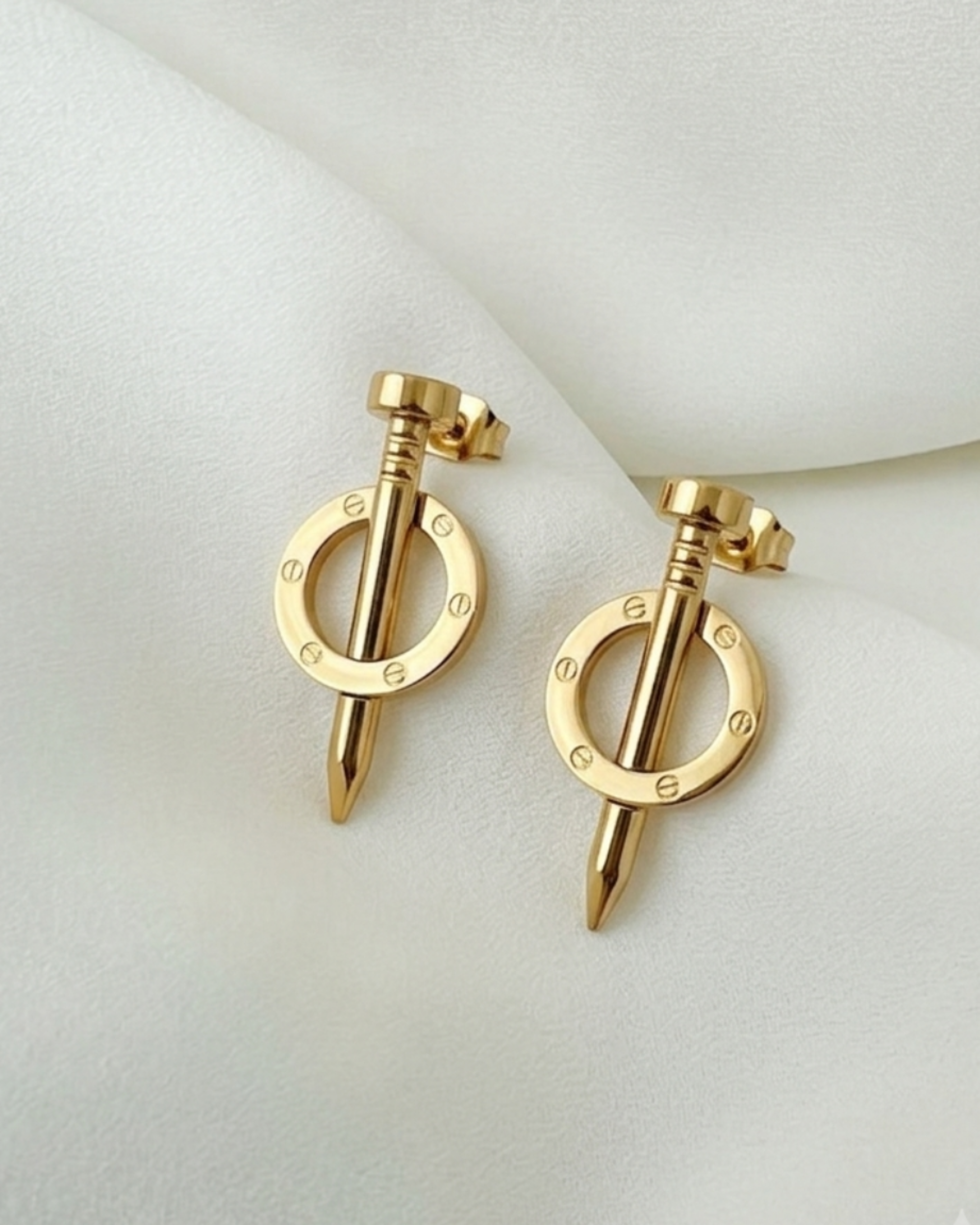 A pair of gold-toned hoop earrings with a central spike and small engraved letters, displayed on a soft white fabric background.