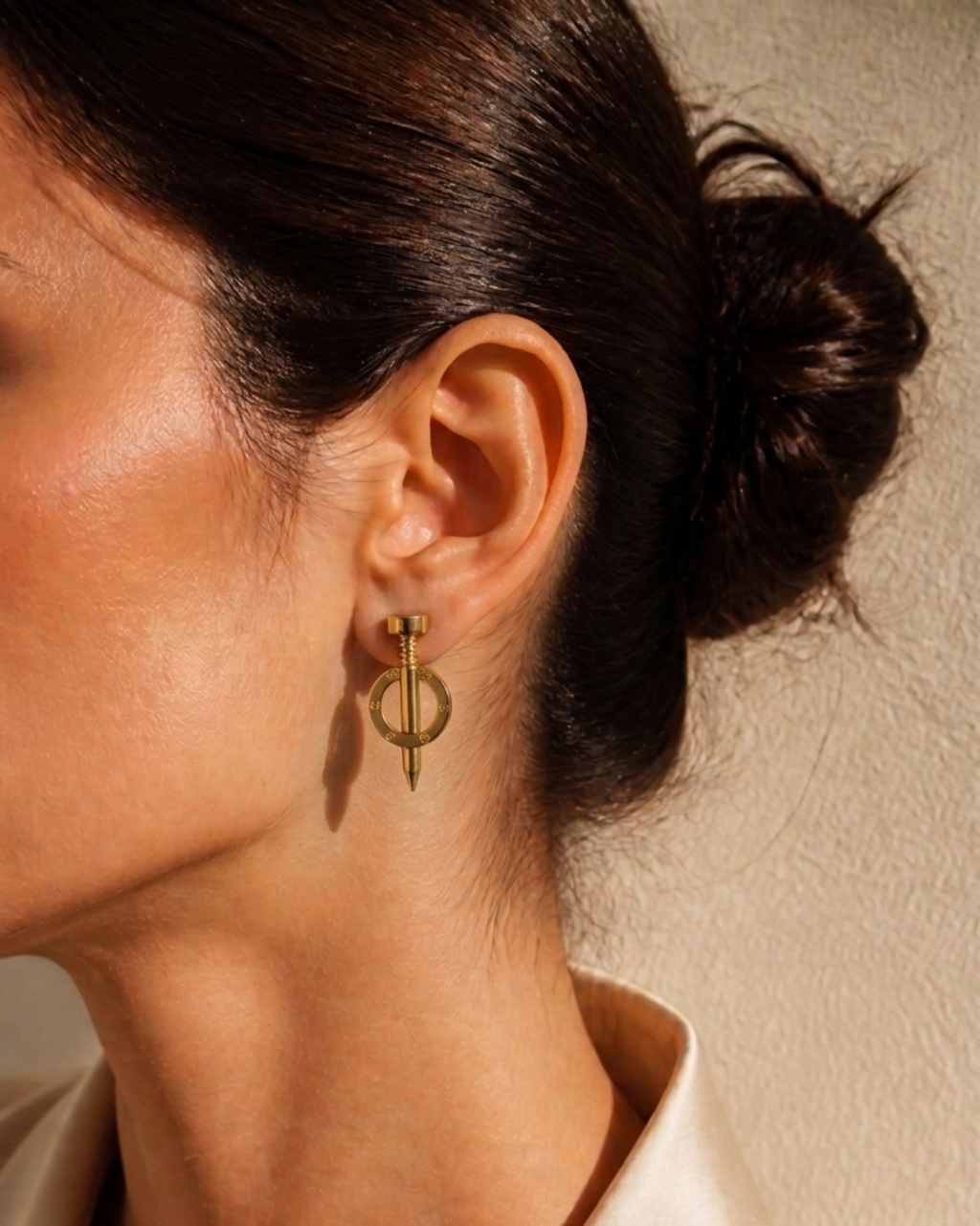 A close-up of a model's ear wearing a delicate gold geometric dangle earring, paired with a cream satin shirt against a textured beige wall.