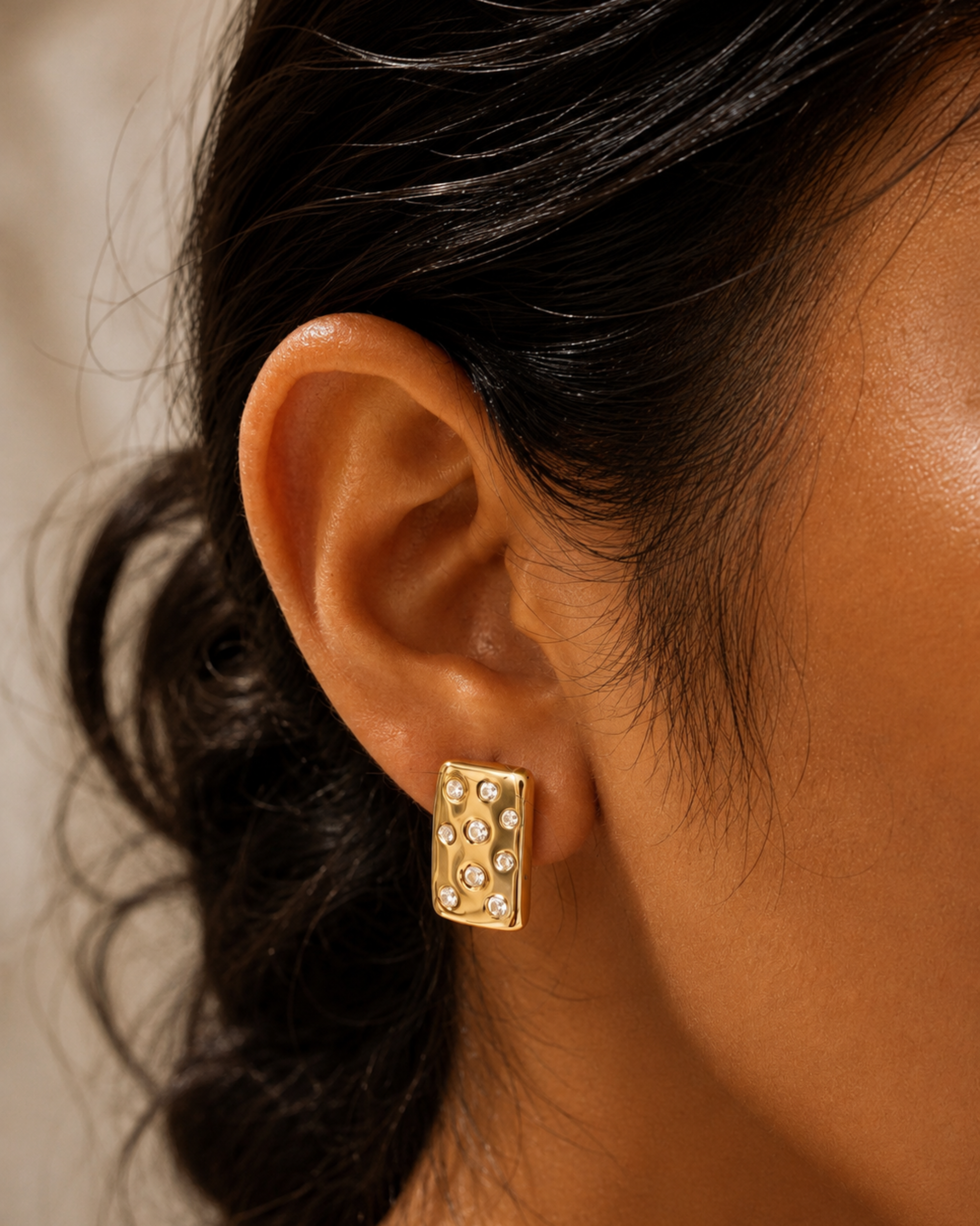 A close-up of a model's ear wearing a gold rectangular stud earring adorned with small clear rhinestones.