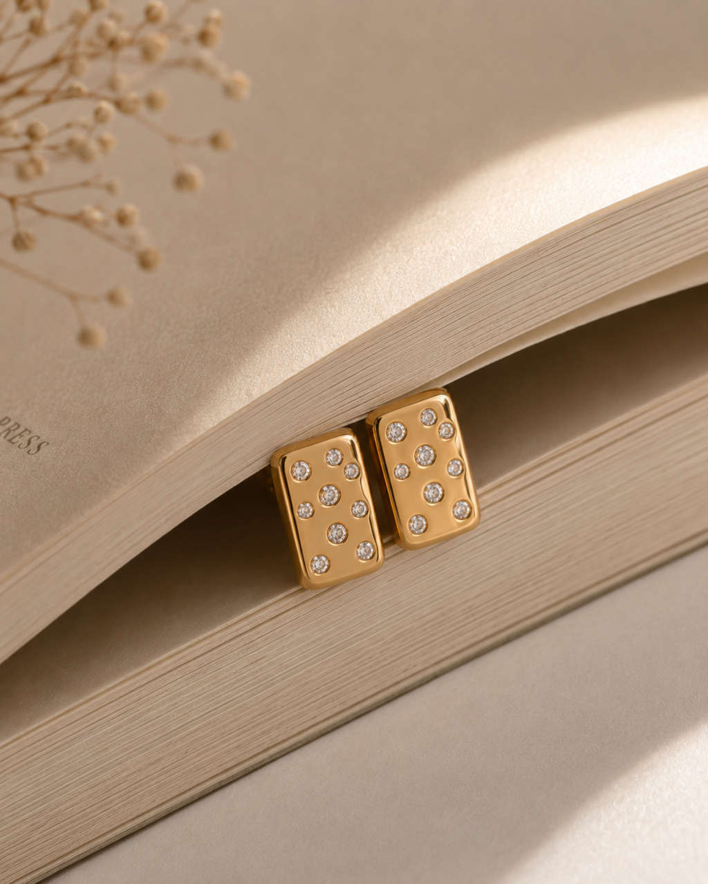 A pair of gold-toned rectangular stud earrings with small clear rhinestones, resting on the pages of an open book with dried floral stems in the background.