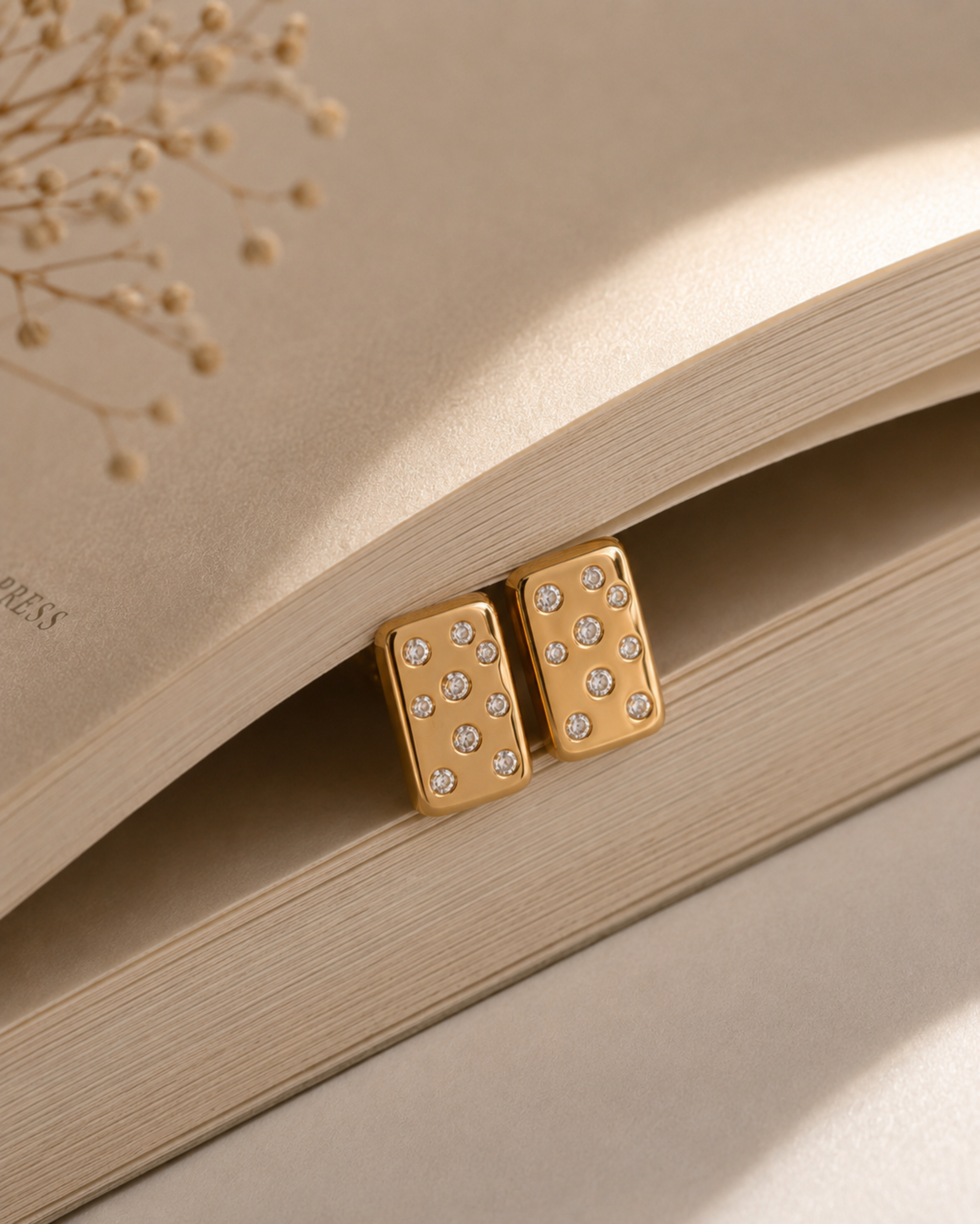 A pair of gold-toned rectangular stud earrings with small clear rhinestones, resting on the pages of an open book with dried floral stems in the background.