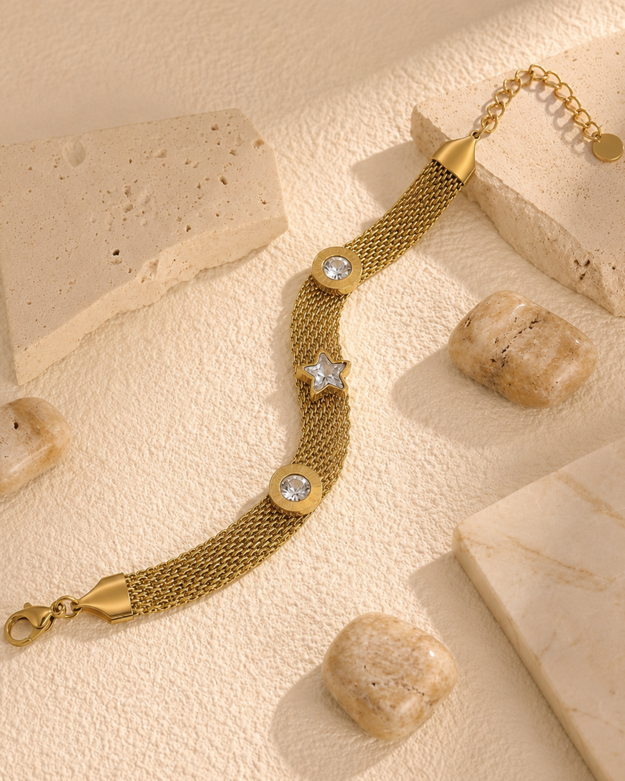 A gold mesh bracelet with a star and oval crystal charms, styled on a textured beige surface with stone props.