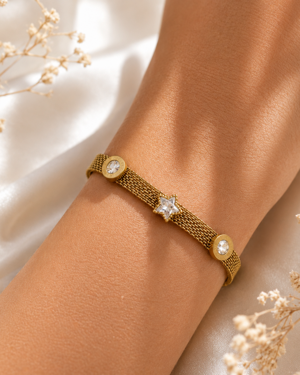 A gold mesh bracelet with a central star-shaped crystal and two round crystal accents, shown worn on a person's wrist against a soft white fabric background with dried flowers.