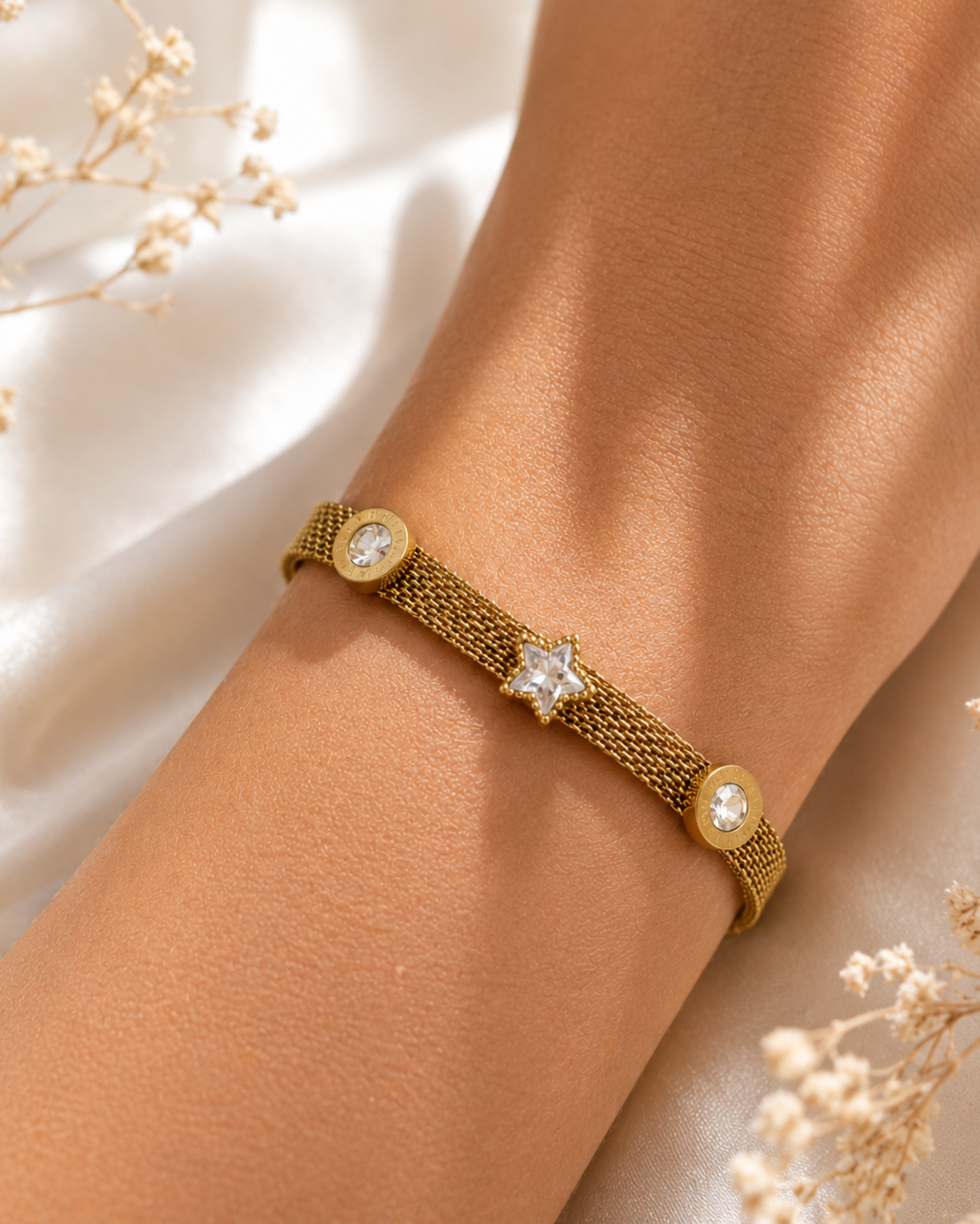 A gold mesh bracelet with a central star-shaped crystal and two round crystal accents, shown worn on a person's wrist against a soft white fabric background with dried flowers.