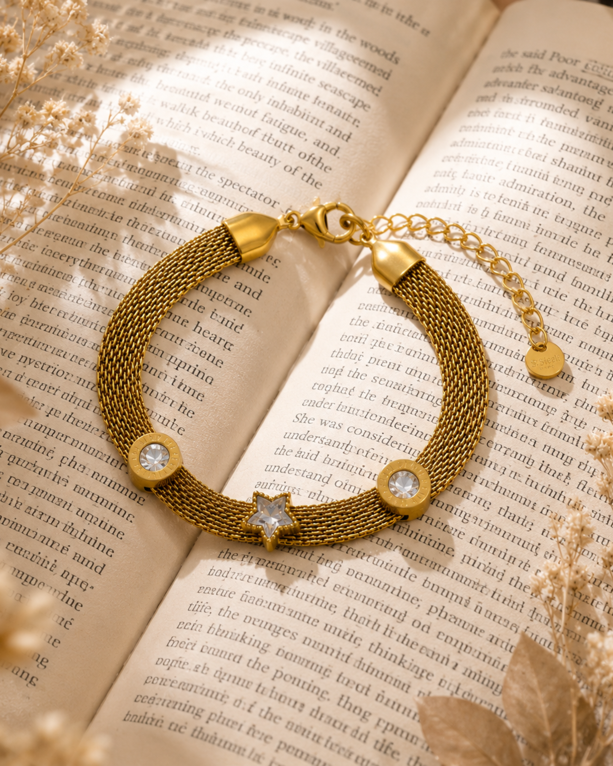 A gold-toned mesh bracelet with three clear crystal-like round charms and a small star charm, displayed on an open book with dried flowers.