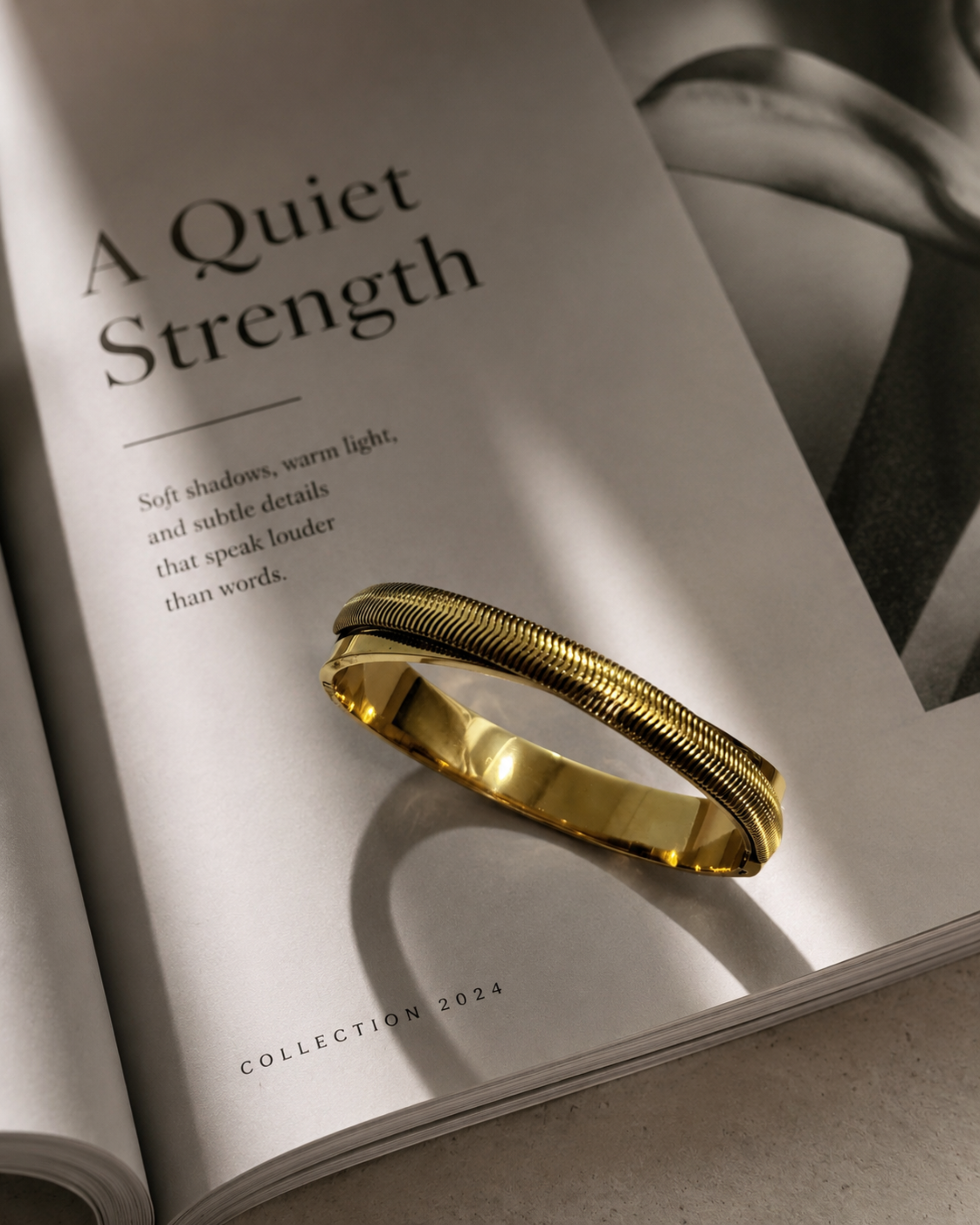 A polished gold bangle bracelet with a textured, ridged outer band and a smooth inner channel, resting on an open book page with the headline 'A Quiet Strength'.