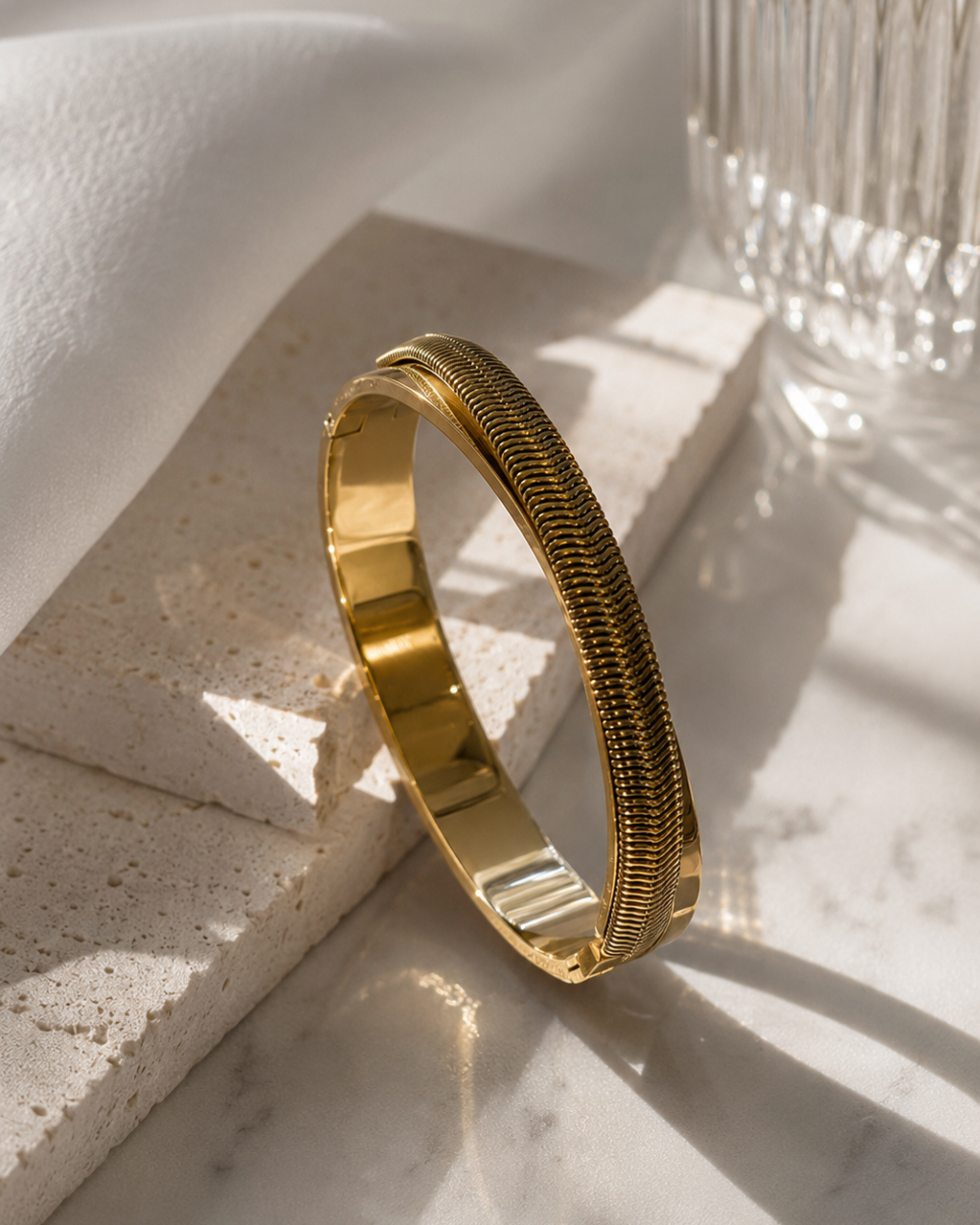 A polished gold bangle bracelet with a textured, ridged outer band, displayed on a light stone block with a glass and draped white fabric in the background.
