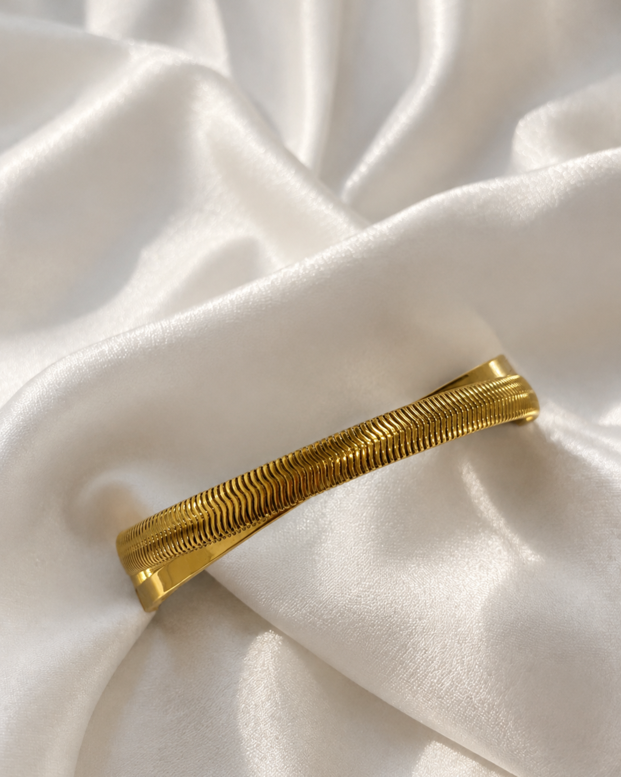 A textured gold bangle bracelet with a ribbed, snake-like pattern, displayed on draped white satin fabric.