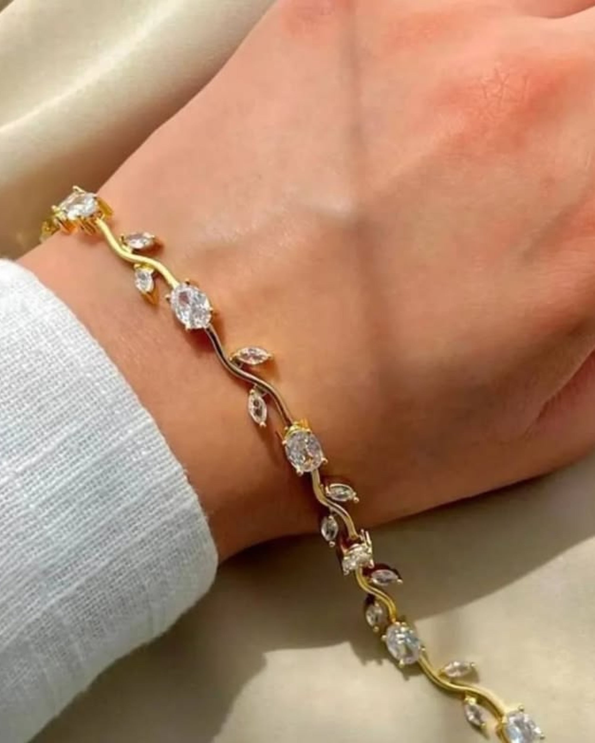 A gold vine bracelet with clear gemstone leaves and small rose gold accents, shown worn on a person's wrist against a soft beige fabric background.