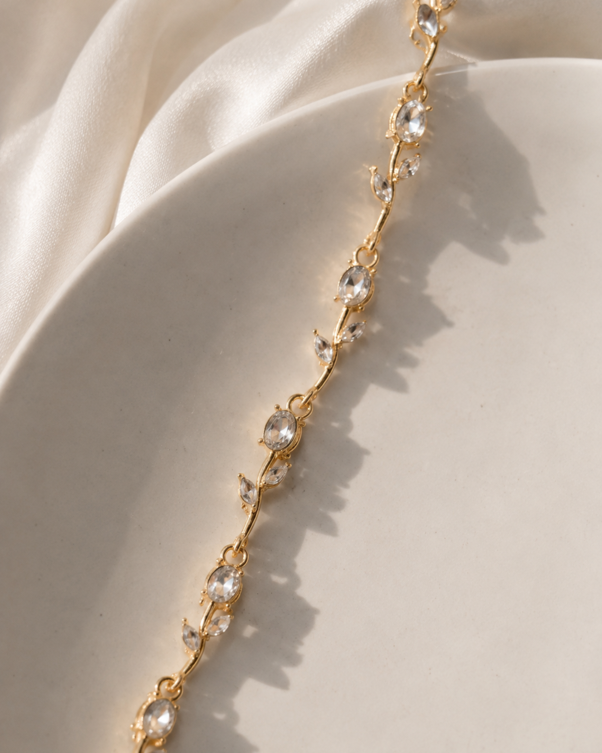 A gold vine-style bracelet with clear teardrop crystals, draped diagonally across a cream satin fabric background.